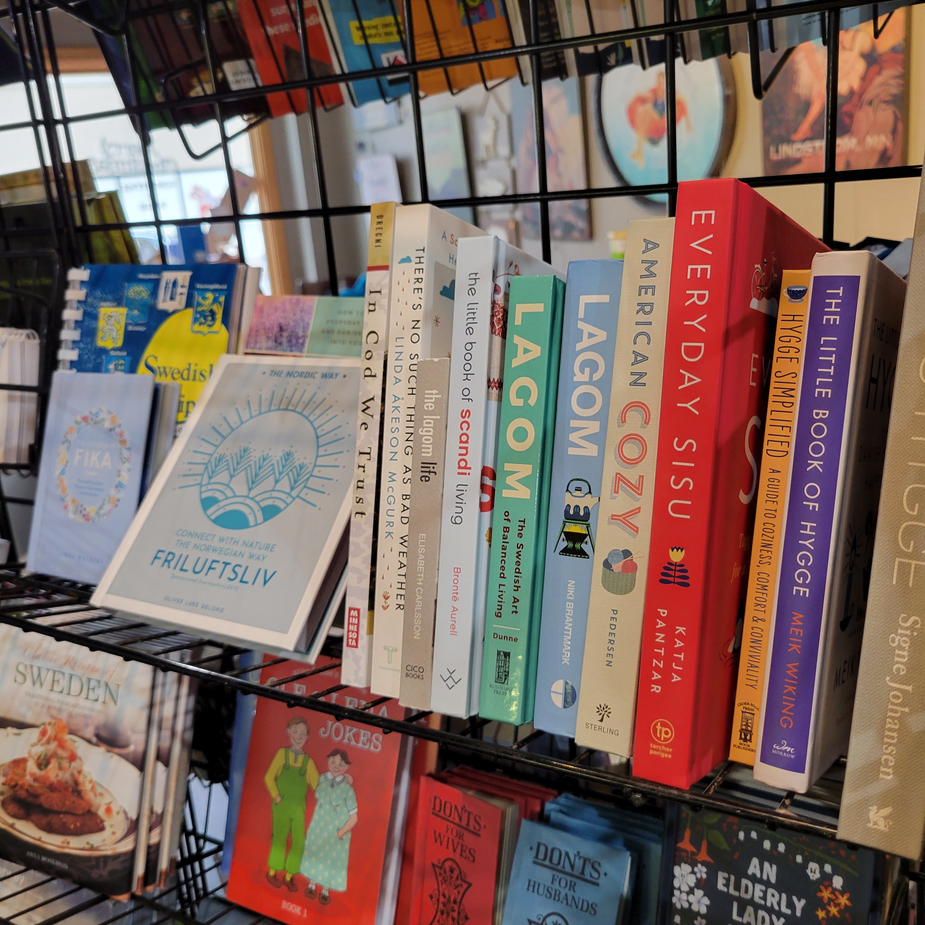 A display of various books and publications on a metal rack, featuring titles on Swedish lifestyle, culture, and self-care, including topics like fika, hygge, and lagom.