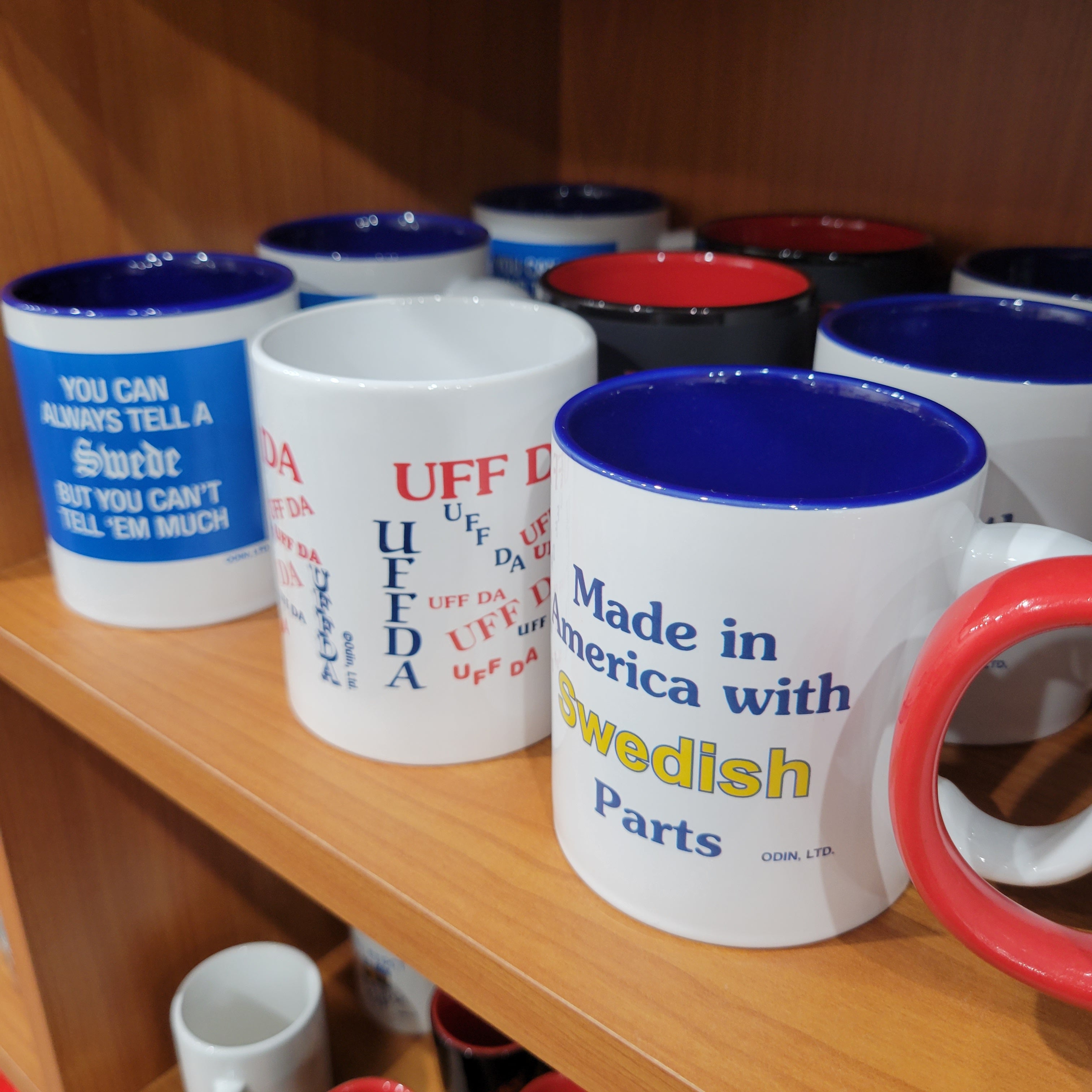 A selection of novelty mugs on a wooden shelf. One mug reads Made in America with Swedish Parts, while others have humorous Swedish-themed phrases.