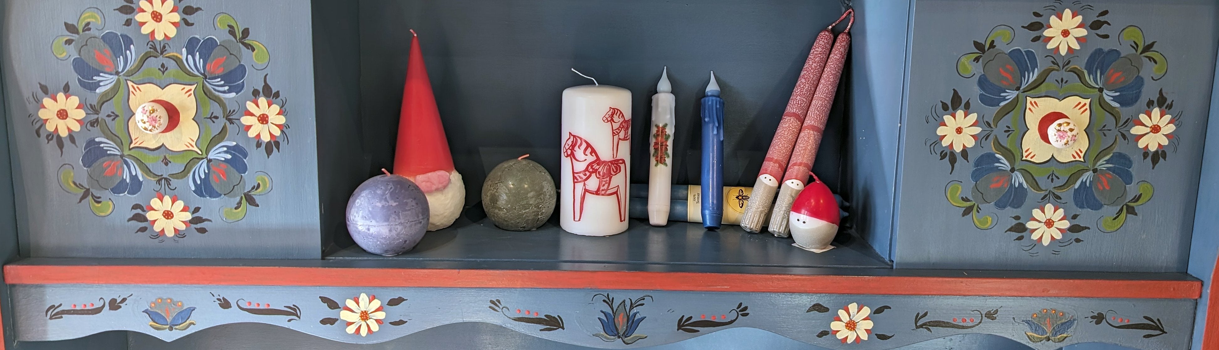 A decorative shelf with colorful floral patterns displays various candles, including spherical, cone-shaped, and pillar styles, alongside a small gnome figurine.