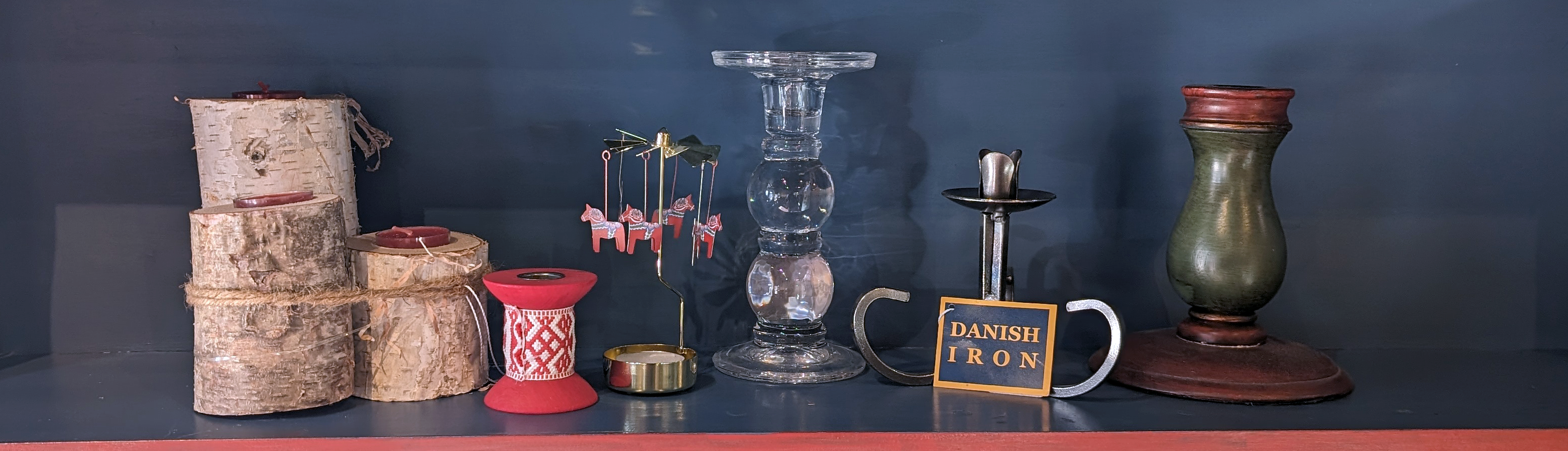 A shelf displays birch logs, red and glass candle holders, a Danish iron sign, and a rotating candle holder with small flags.