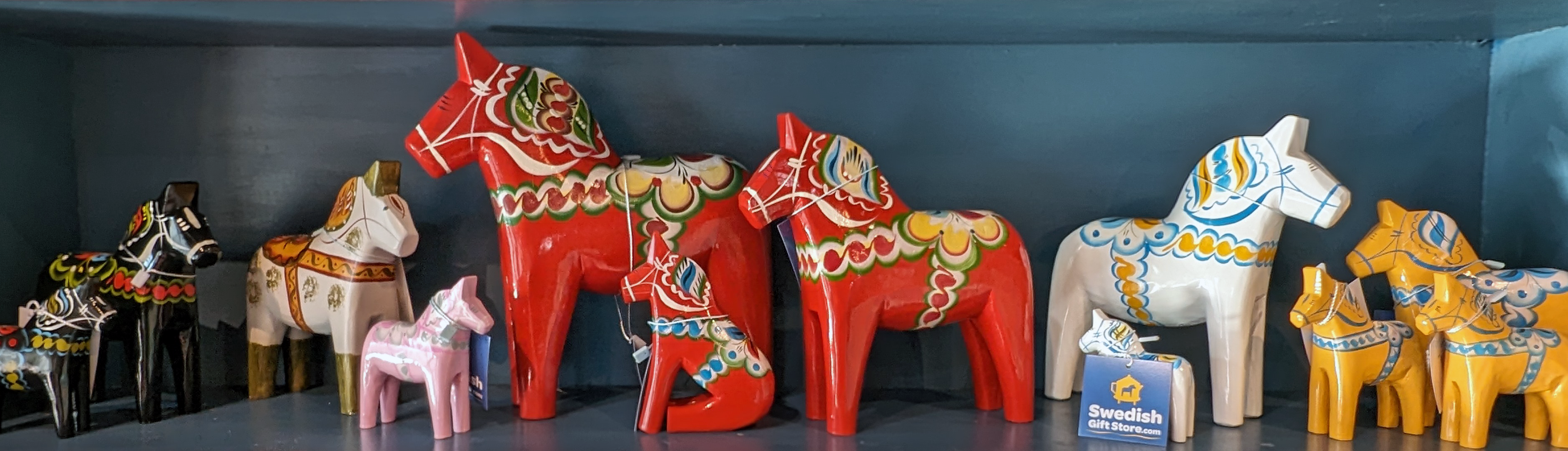 A display of colorful painted wooden Dala horses in various sizes, featuring traditional Swedish patterns and vivid colors, arranged on blue shelves.