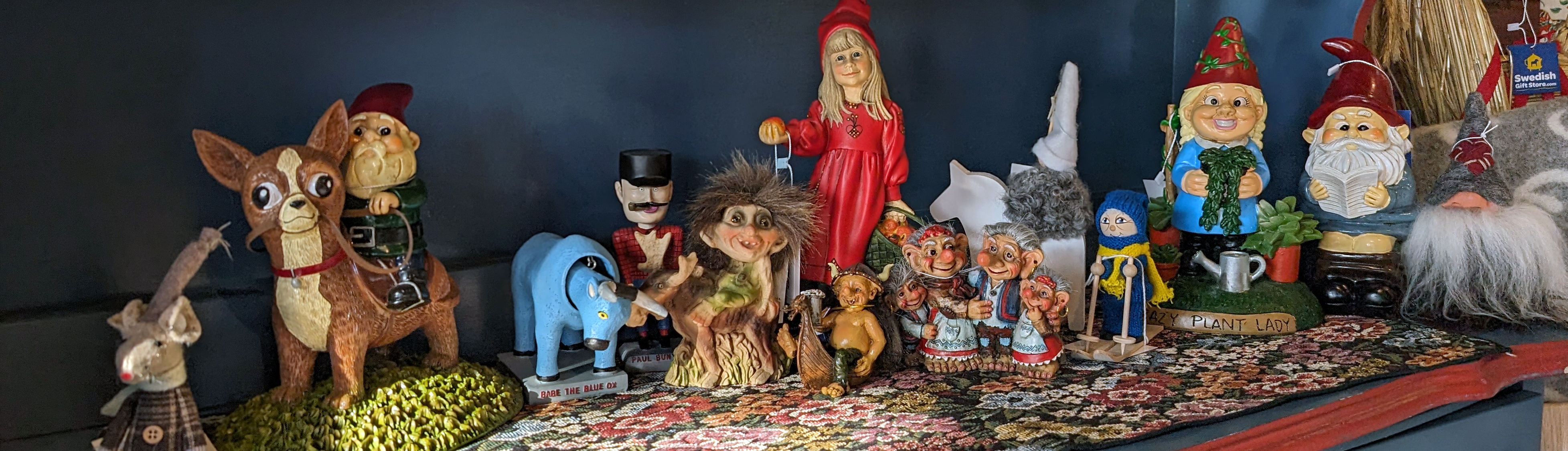 A collection of various whimsical figurines, including gnomes, elves, and animals, arranged on a floral-patterned shelf.
