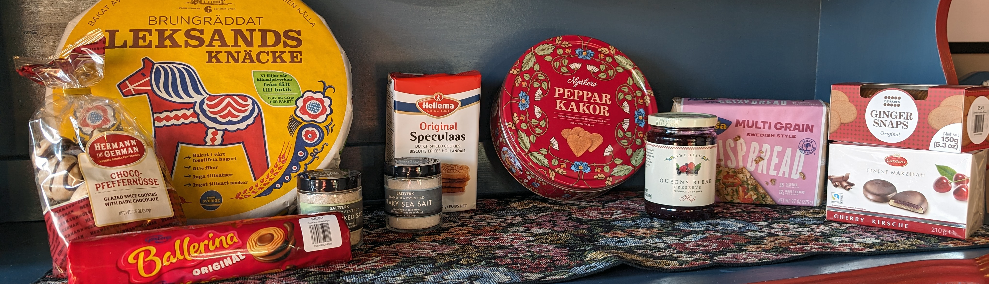 A selection of Scandinavian food items, including crispbread, cookies, ginger snaps, seasoning jars, lingonberry jam, and chocolates, arranged on a shelf.