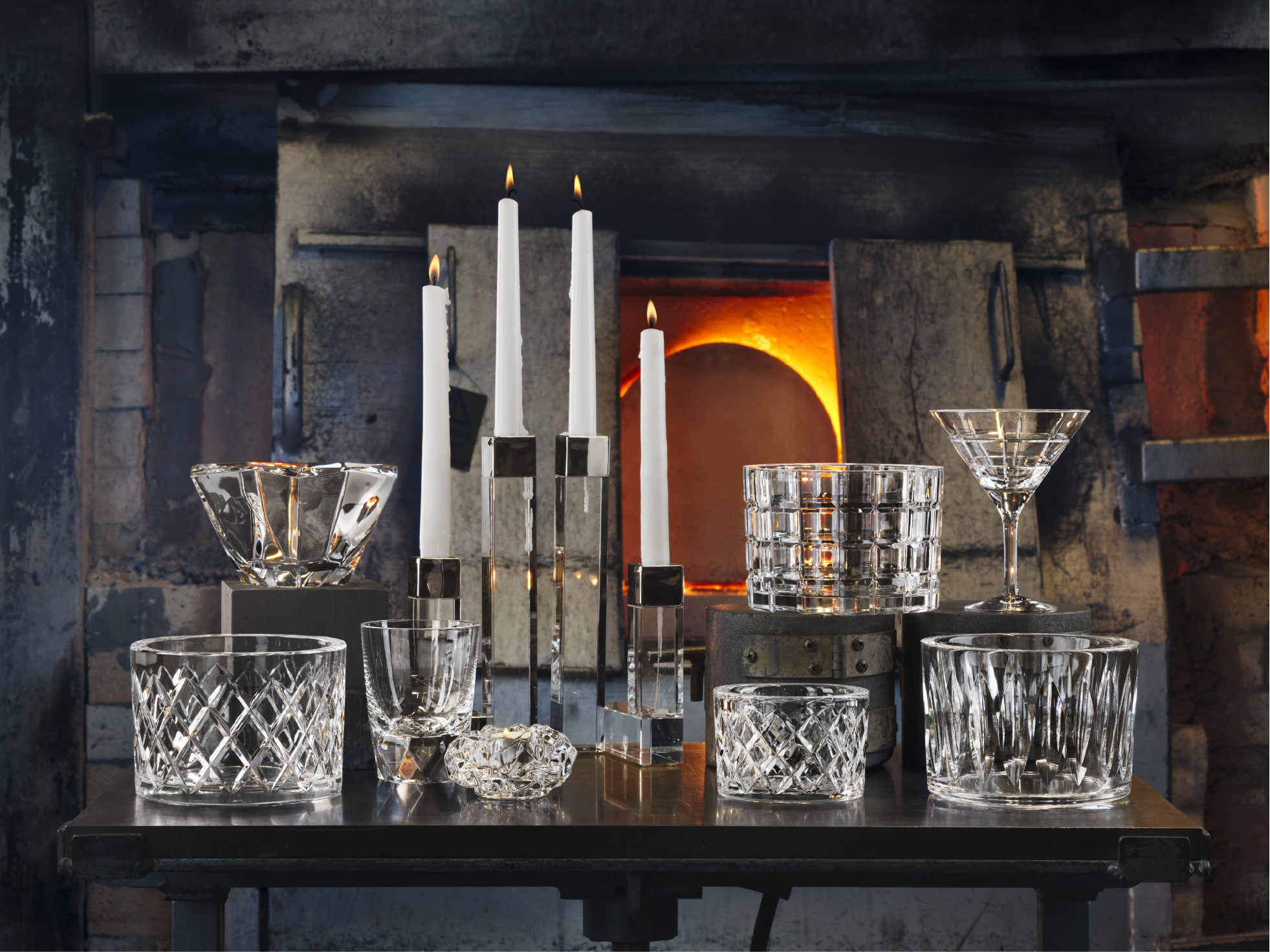 Assorted crystal glassware and candle holders are arranged on a table in front of a lit furnace in what appears to be a glassblowing studio.