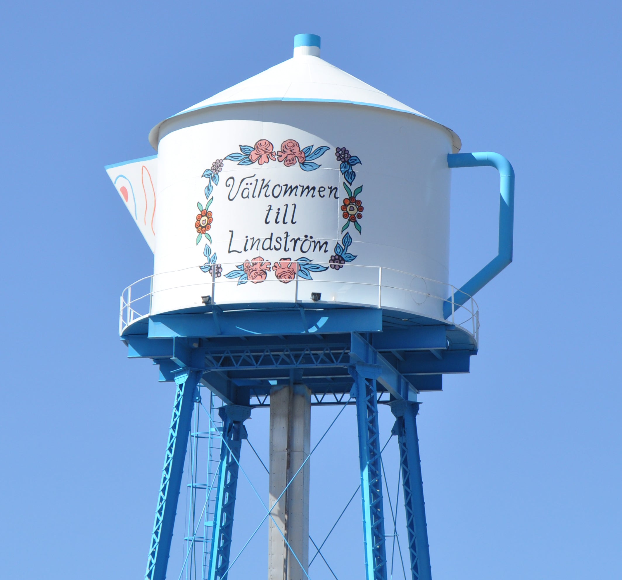 Water tower shaped like a teapot with the text Välkommen till Lindström and decorative flowers, set against a clear blue sky.