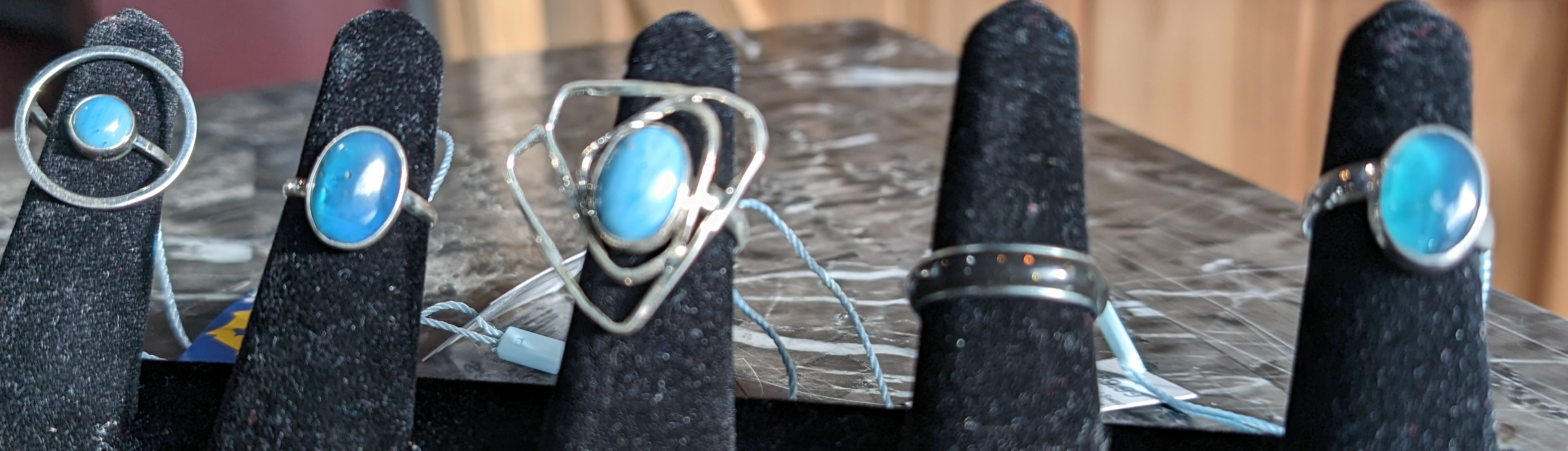 A display of five rings with blue stones set in silver, each on a black stand against a blurred background.