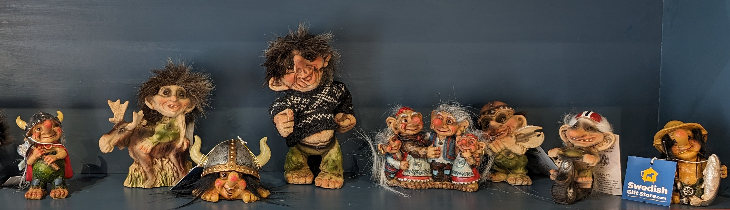 A collection of troll figurines displayed on a shelf, including a Viking helmet, various hair and attire styles, and a small card labeled Swedish Gift Store.