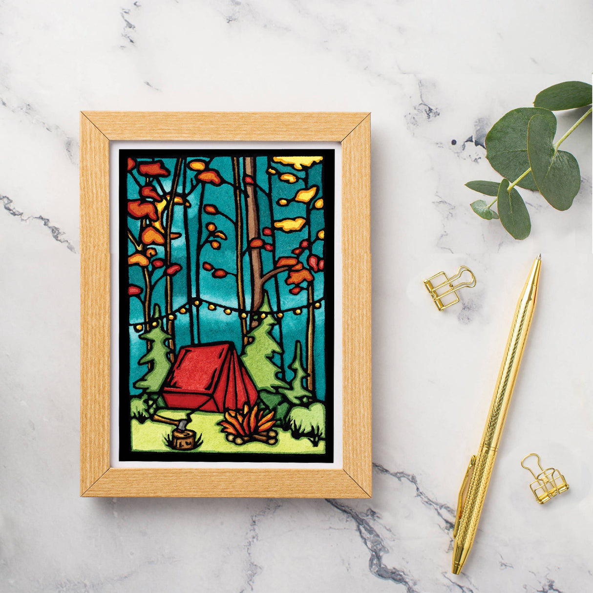 A greeting card titled "Evening at Camp Tent" by Sarah Angst features artwork of a red tent in a forest with orange leaves and a campfire, elegantly displayed on a marble surface alongside a pen and paperclips.