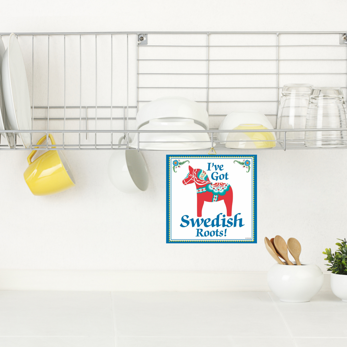 A dish rack with plates, bowls, cups, and glasses sits above a counter with utensils; a 6x6 Ive Got Swedish Roots! Swedish Dala Horse Wall Plaque decorates the wall, celebrating Swedish heritage.