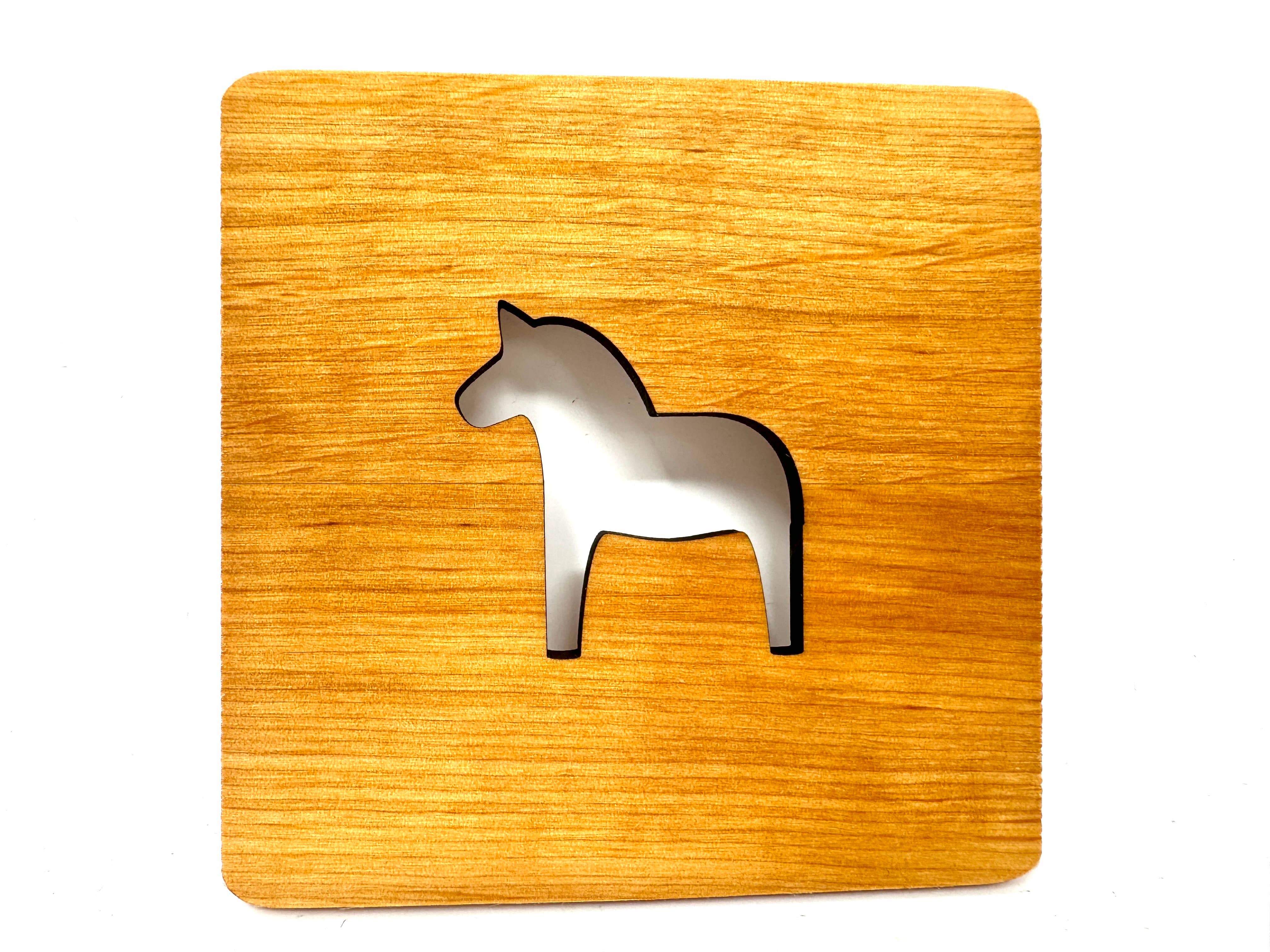 A charming square coaster, laser cut from alder wood, features a silver Swedish Dala Horse silhouette at its center.
