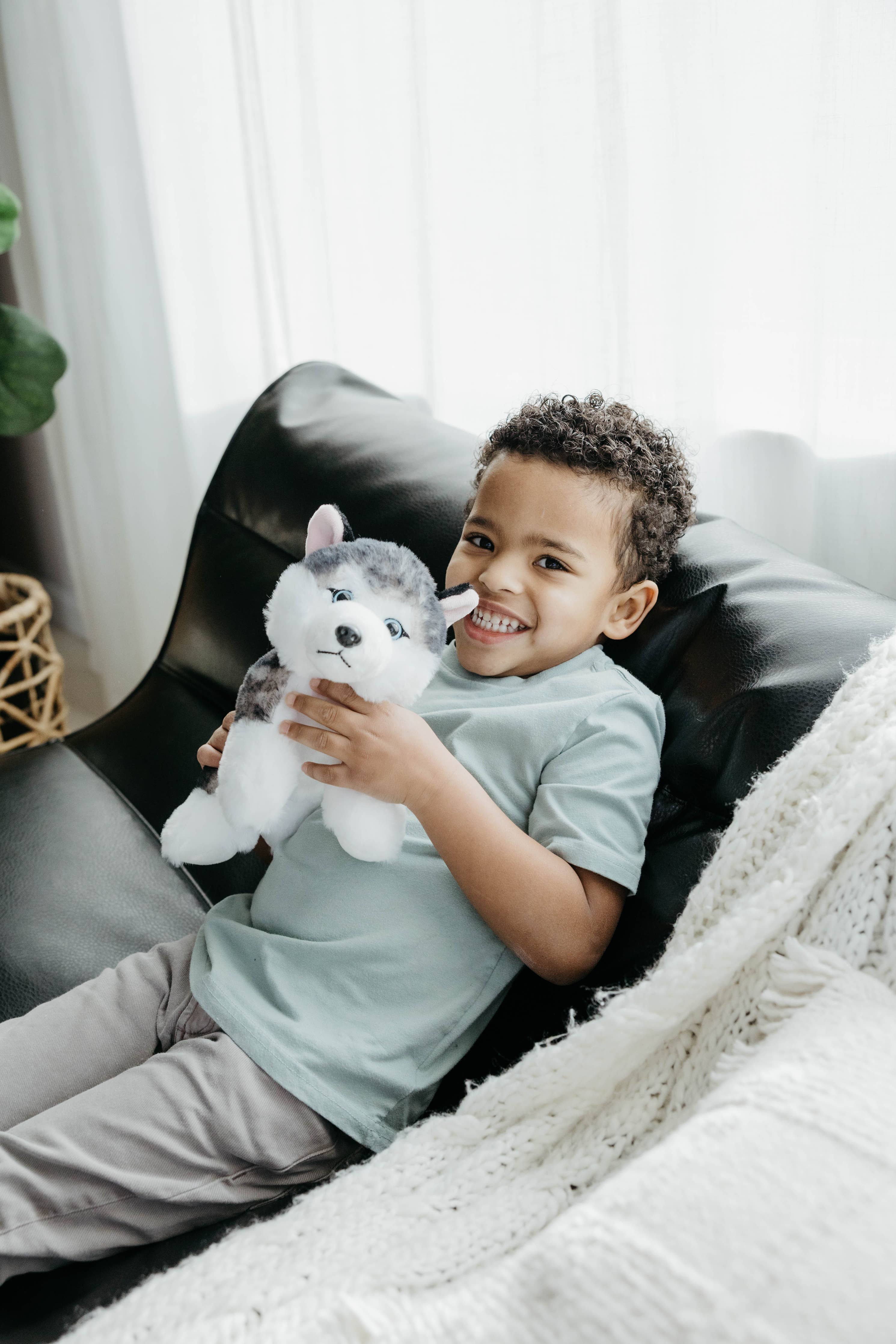 A young boy smiles as he sits on a black sofa, holding the Plush: Husky Dog 9 Tall Stuffed Animal.