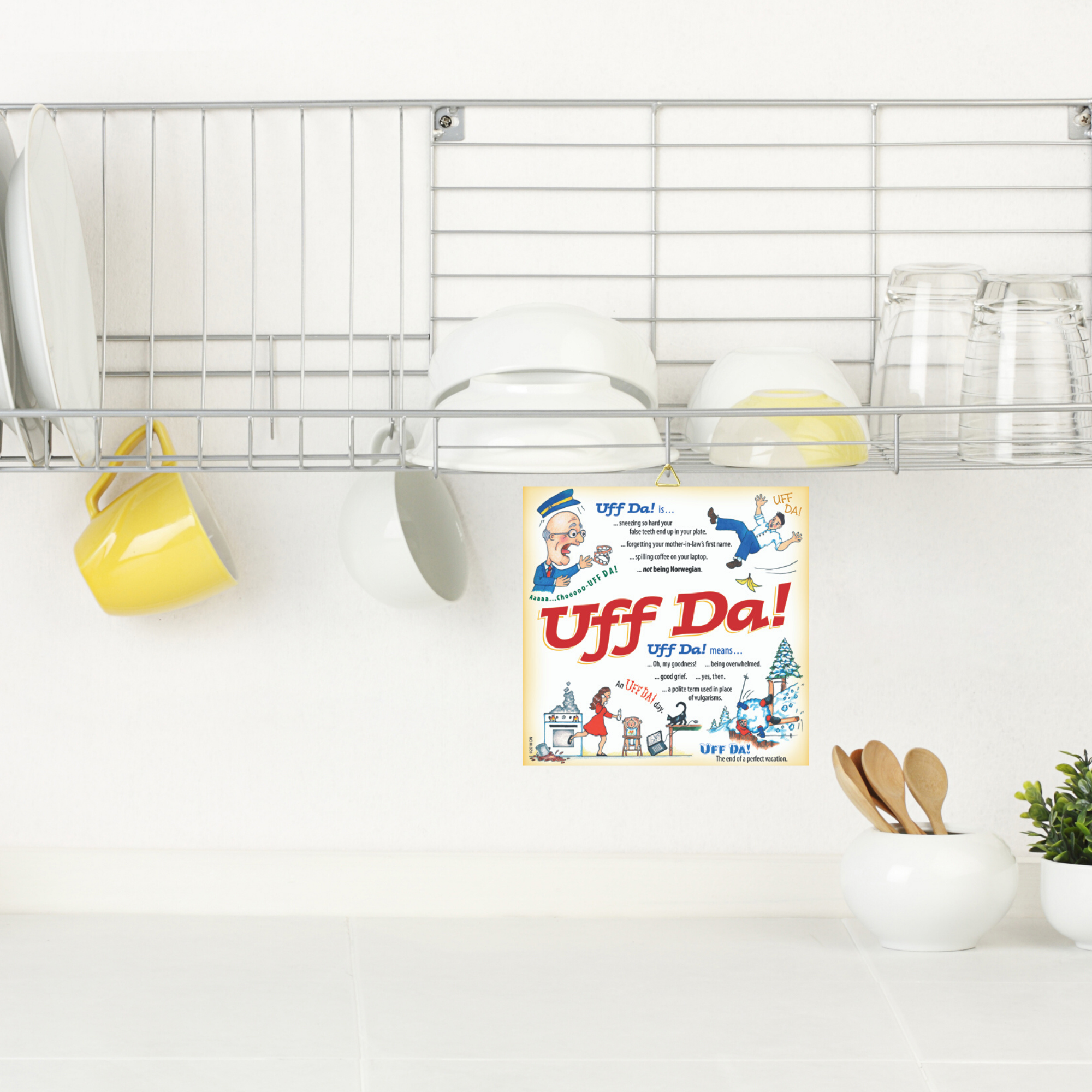 A kitchen dish rack with white dishes, bowls, glasses, and three mugs sits above a Scandinavian Gift Uff Da Ceramic Wall Tile featuring a cork back and hook for hanging.
