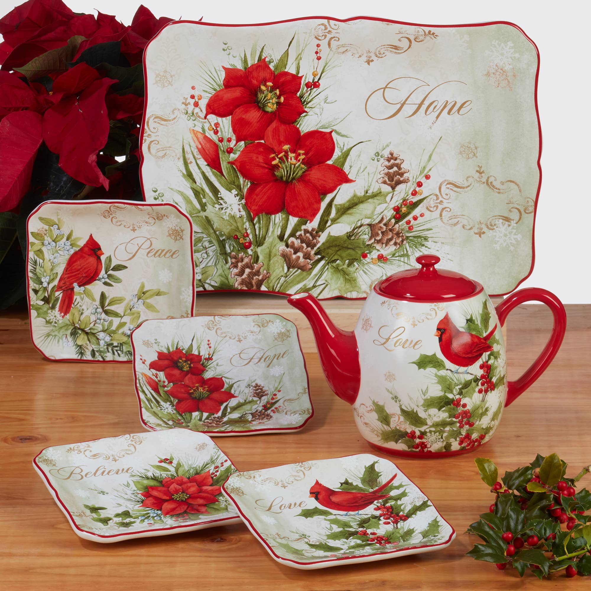 A festive tea set featuring the Winters Medley Cardinal Teapot 40 oz., decorated with red cardinals, poinsettias, and words like Hope, Peace, Believe, and Love, displayed on a wooden table.