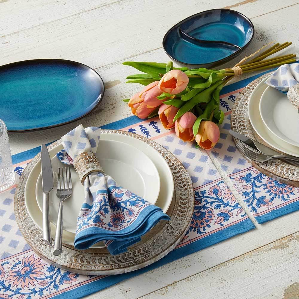 A table set with patterned plates, the Lake Life Ceramic Divided Oval Dish, silverware, blue napkins, a glass, a floral table runner, and a bouquet of pink tulips on a white wooden surface.