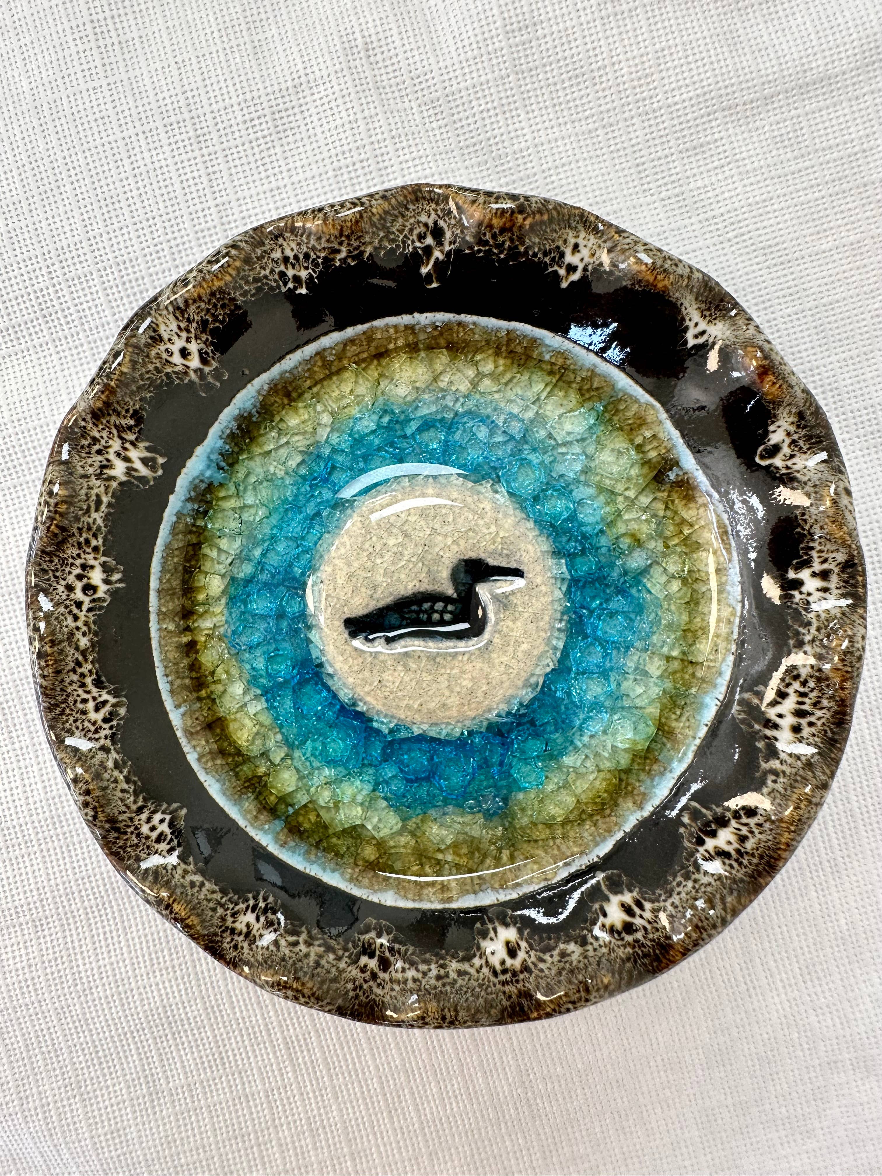 The Dish: Loon Handmade Pottery Dish features recycled glass and clay, a textured rim, blue and green glazed center, and a black loon silhouette. It measures 4.5" in diameter and is displayed on a white cloth background.