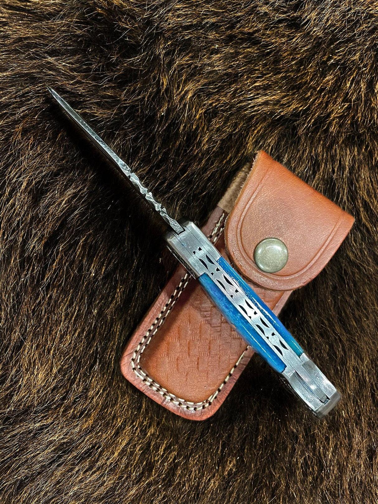 The Knife: Dyed Blue Bone 6.5 Damascus Steel, featuring a patterned blade and blue handle, rests on a brown leather sheath atop textured fur.