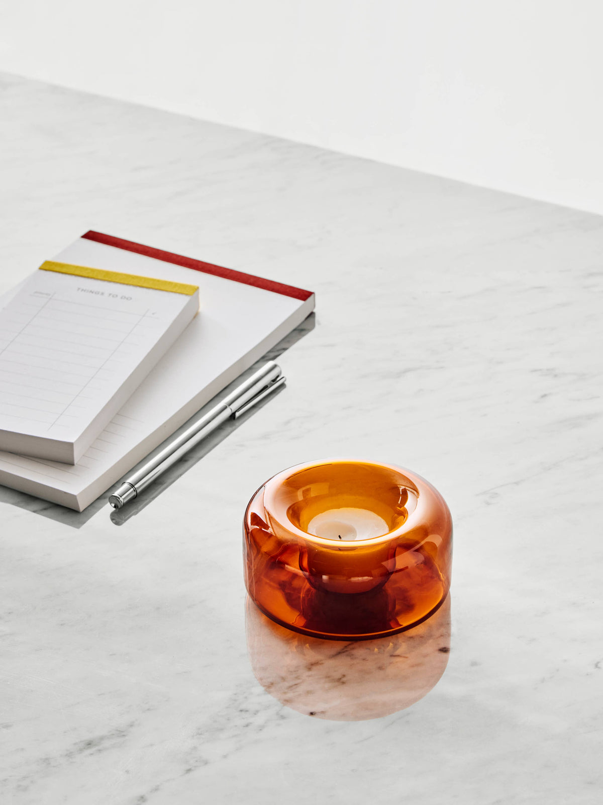 Amber blown glass votive candle holder on a white marble surface, positioned next to a stack of notepads and a silver pen.