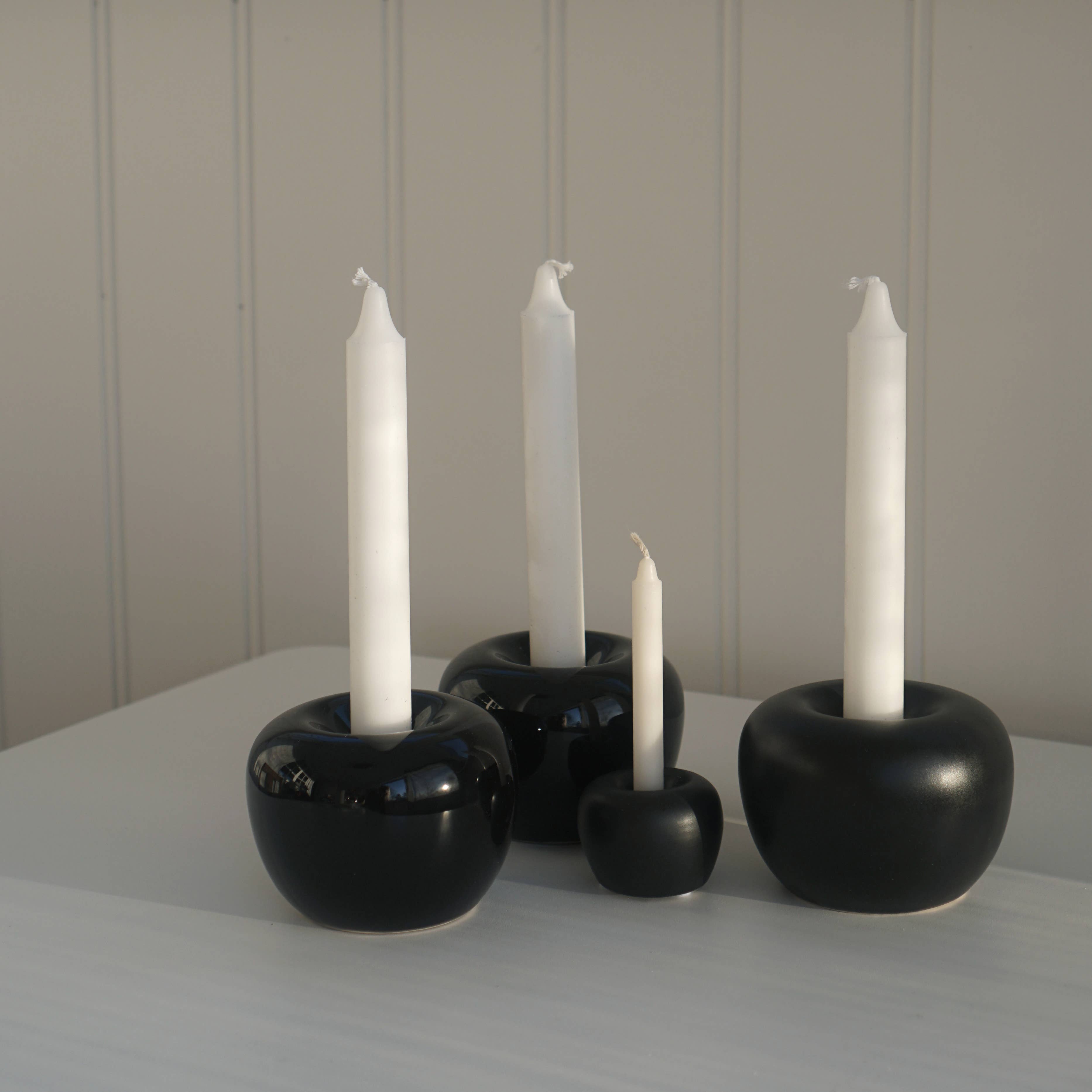 Four white candles of varying heights sit elegantly in black, round stoneware holders on a white surface against a paneled wall, evoking the refined aesthetic of Candle Holder: Apple Candleholder 2-Pack Mini Apple.
