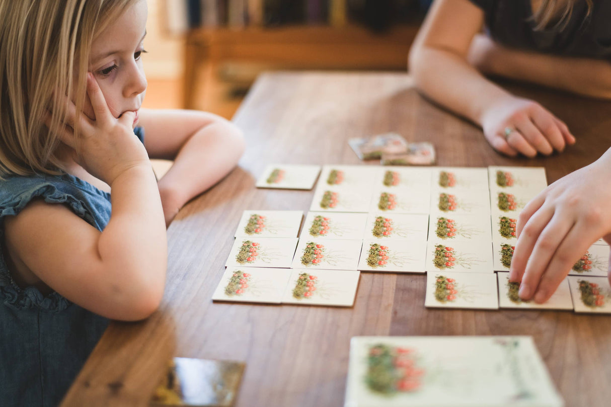 An adult and child play the Elsa Beskow Children of the Forest Memory Game with illustrated cards laid out on a wooden table. The child, looking thoughtful, rests her chin on her hand as they search for matching pairs.
