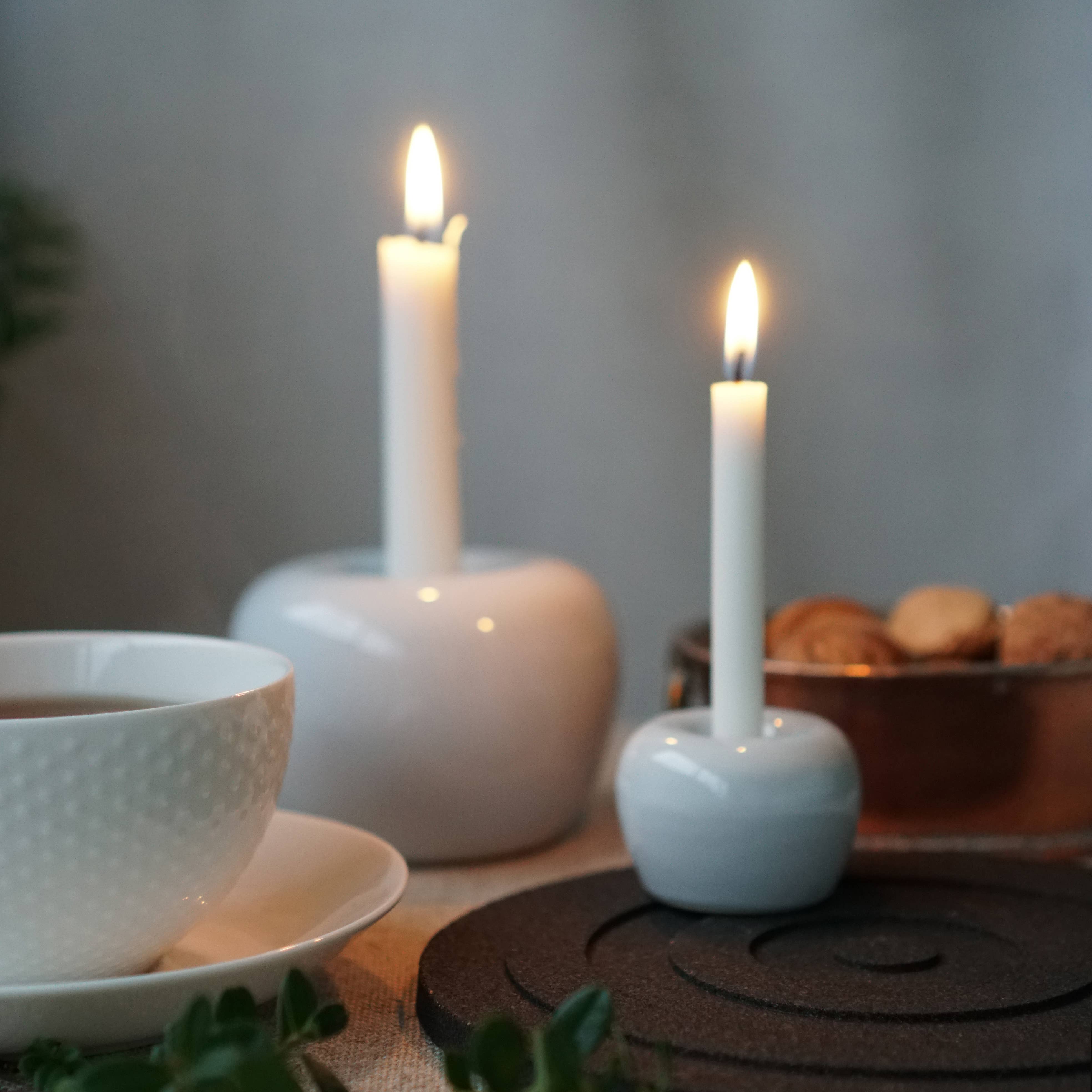 Two lit white candles in Kim Vejsholts Apple Candleholder 2-Pack Mini Apple sit beside a teacup and saucer. A dark coaster and a bowl of nuts add depth to this cozy setting.