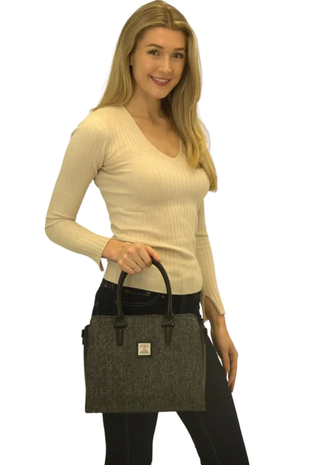 A woman in a beige ribbed long-sleeve top and dark pants holds the Harris Tweed Double Handle Tote in grey against a white background.