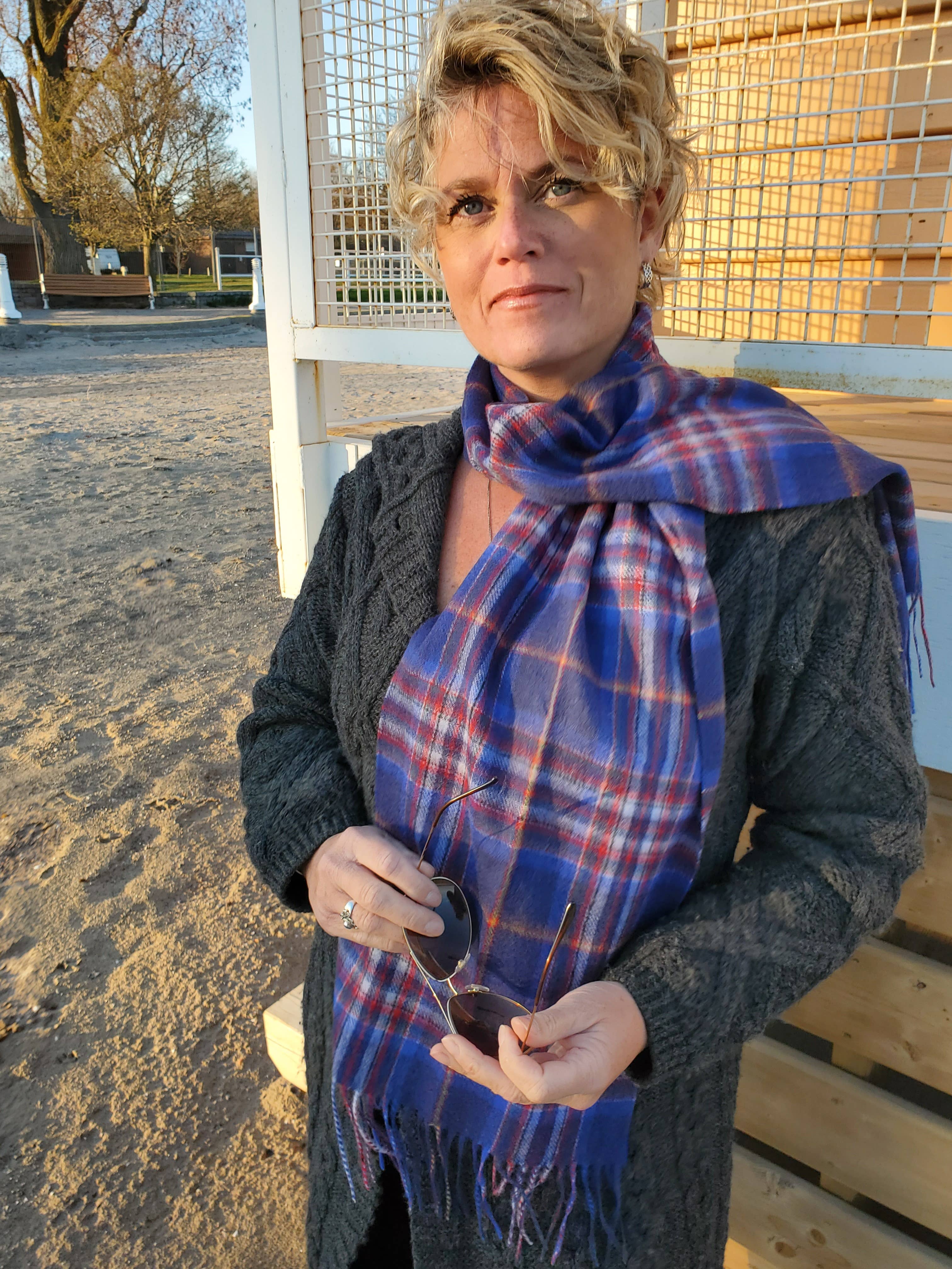 A woman stands outdoors on sandy ground by a wooden structure, holding sunglasses and wearing a gray sweater and the Esprit du Quebec Lambswool Scarf, sunlight illuminating her face.