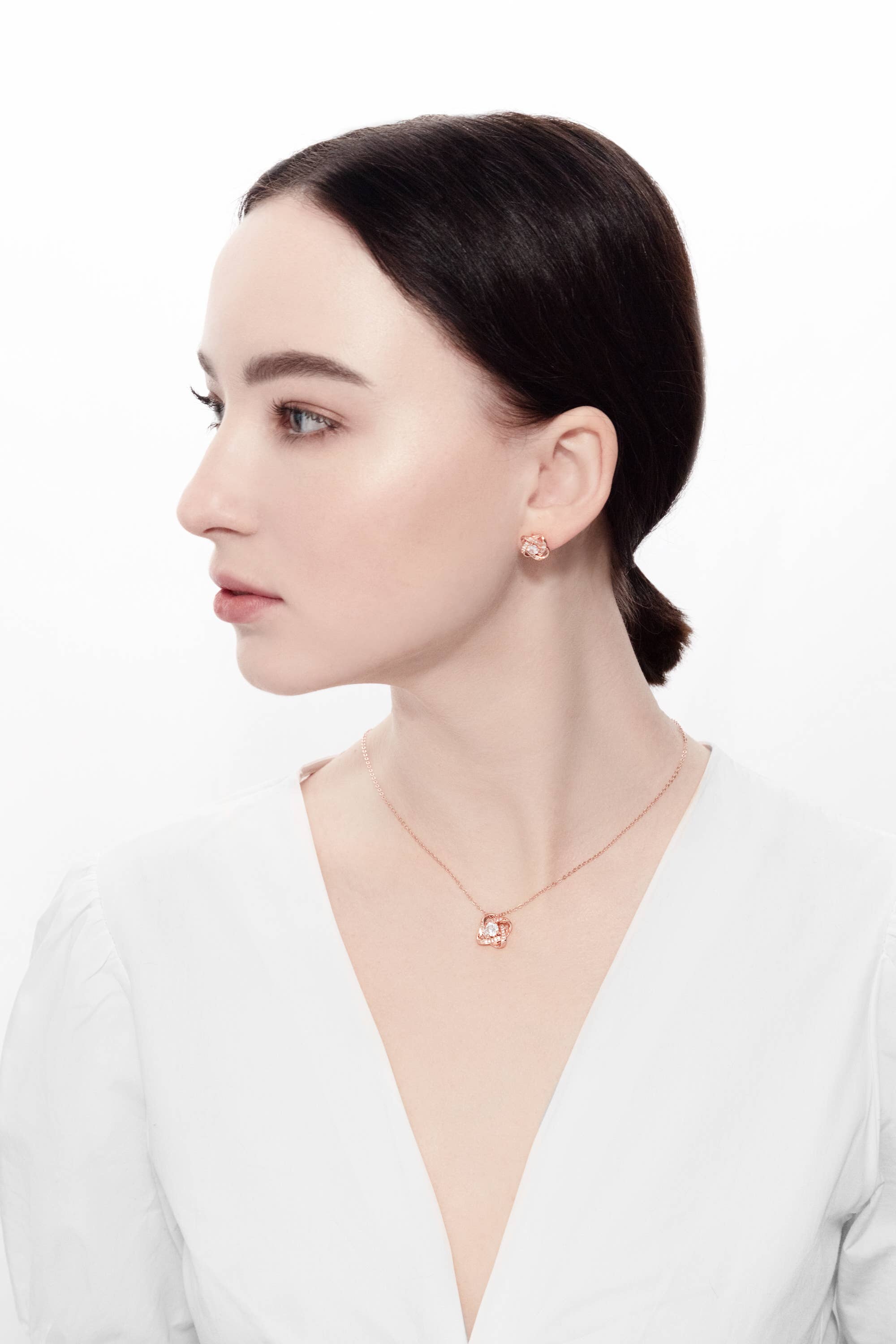 A woman with dark hair in a low bun wears a white top, rose gold earrings, and the Ice Dance Rose Gold/Sterling Silver Necklace with Brilliant Cut Cubic Zirconia, looking to the side against a plain white background.
