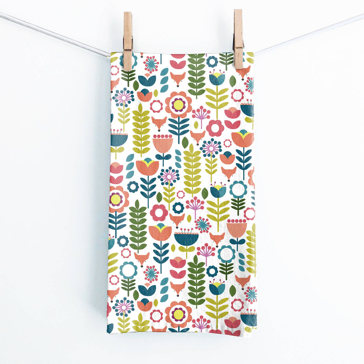 The Juniper Blue "Tuli" Scandinavian Floral Folk-Art Tea Towel (19x27 in) with hanging tab, featuring bright stylized flowers and leaves, hangs from a white clothesline with two wooden clothespins against a plain background.