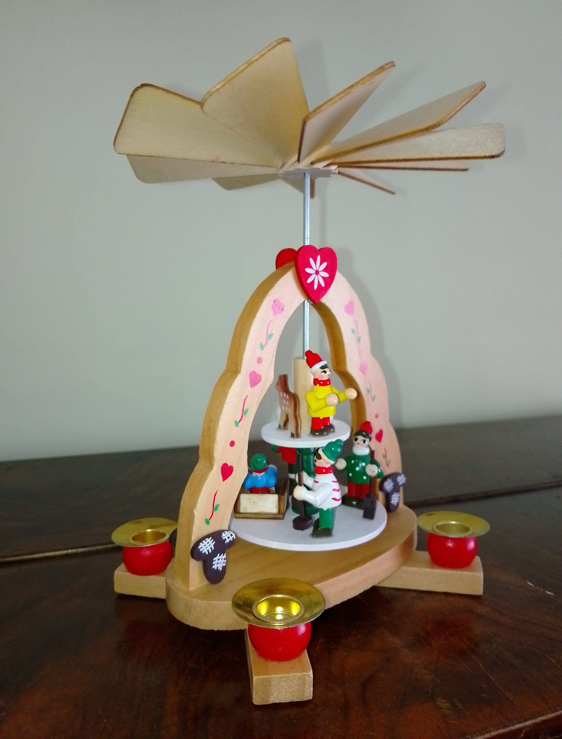 The "Kids" Pyramid Candle Holder is handcrafted in Germany, stands 7" tall by 6" wide, holds 4 candles, and features figurines with a spinning fan top, displayed on a dark wooden surface.
