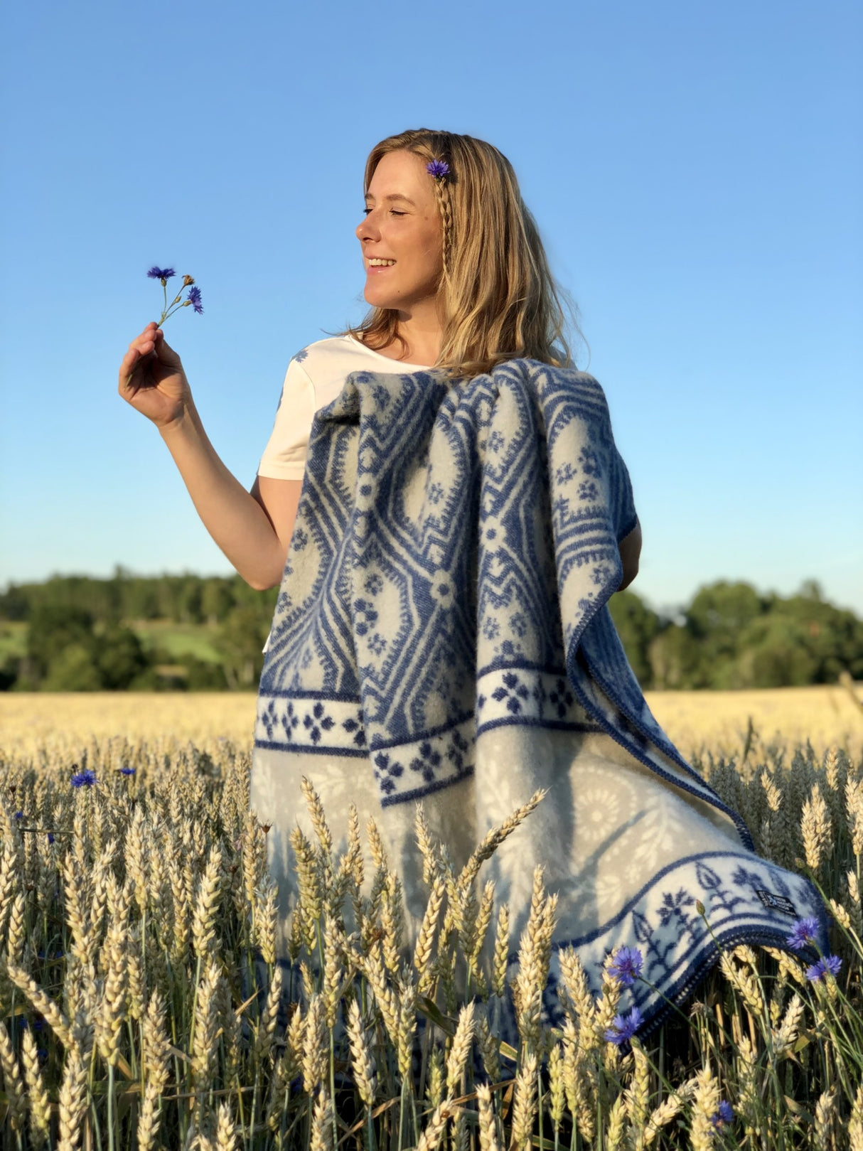 In a peaceful wheat field, a woman wrapped in the Blaklint Cornflower blanket—crafted from 100% Norwegian lambs wool by Kerstin Landstrom—holds a flower, presenting a scene of blue and white harmony.