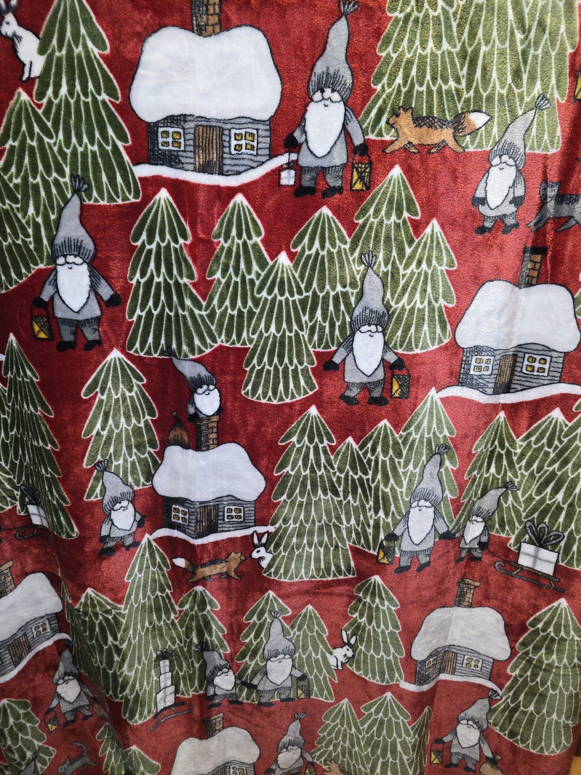 The Tomtehus Fleece Throw Blanket (51 x 67) features snow-covered trees, small houses, and gnome-like figures with lanterns on a red background—perfect for adding cozy Swedish charm to your home.
