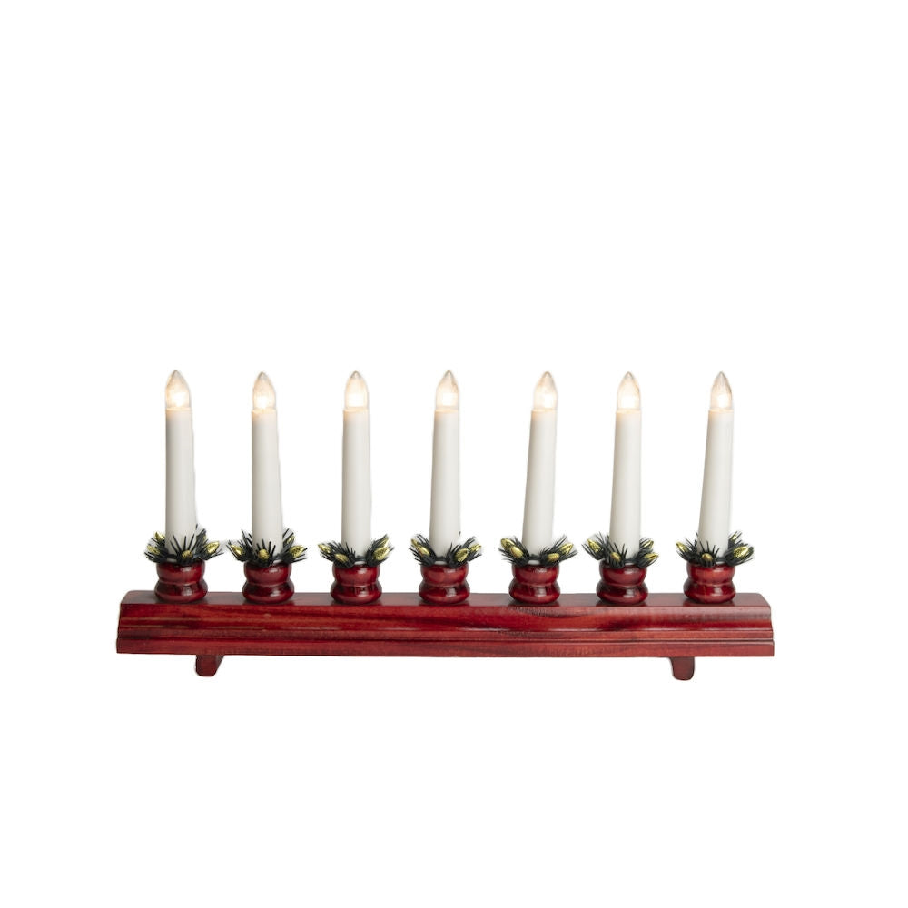 The Konstsmide - Ingrid, an electric 7-light candelabra, showcases a row of beautifully crafted white candles with glowing bulbs in red holders, sitting elegantly on a red stained wooden base and adorned with green garland.