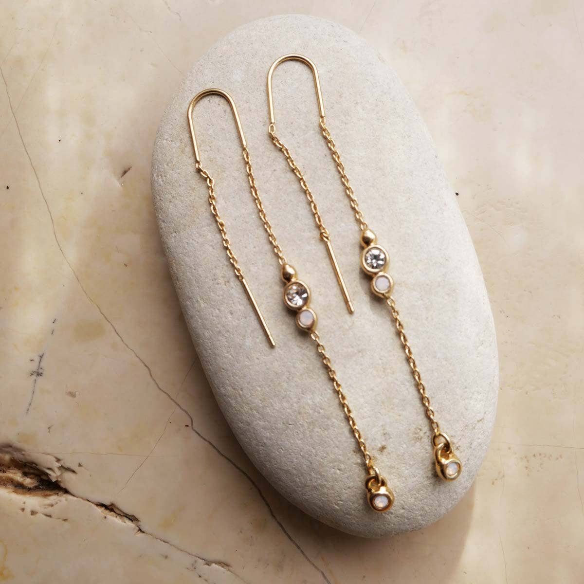 The A&C Oslo Long Crystal Chain Gold Earrings, featuring small round white stones and clear gems, are displayed on a smooth oval stone against a marble background, highlighting the elegance of organic jewelry design.