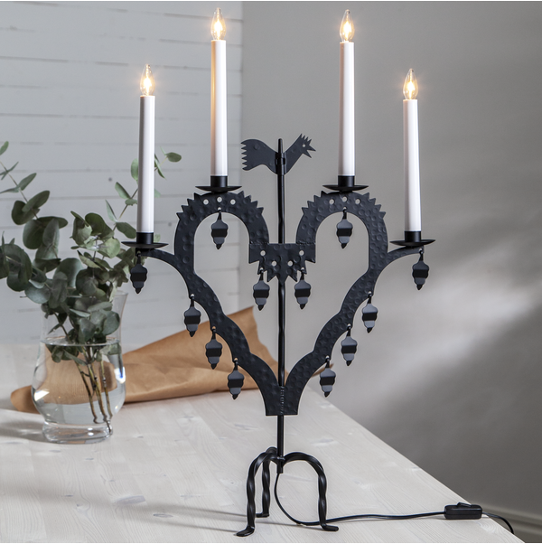 The Star Trading Metal Candlestick El-Tupp, a black heart-shaped candelabra with four lit candles, stands on a light table beside a glass vase with green foliage, creating a cozy Christmas atmosphere.