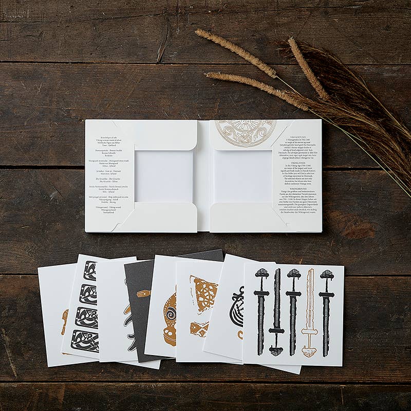 On a dark wooden surface, an open white envelope displays decorative cards with intricate black and brown designs, complemented by tall dried grass. The scene evokes subtle Viking motifs and a nod to Danish history. Product: Card Pack - Viking Finds Square Card Folder by Koustrup & Co, Denmark.