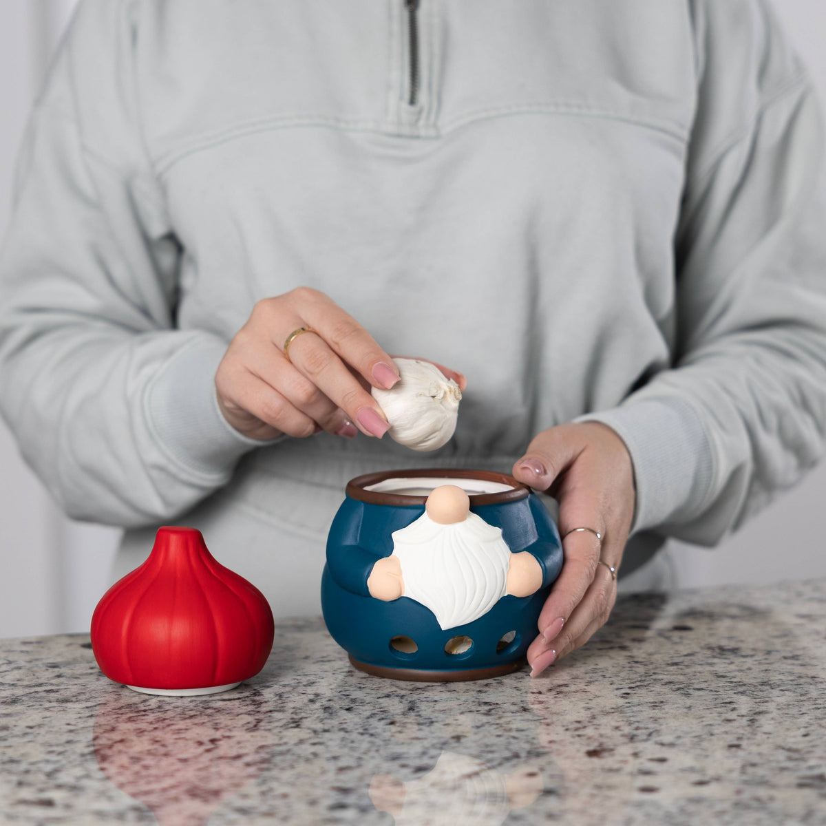 A person puts garlic into a blue gnome-shaped garlic keeper on a granite countertop with a red, garlic-shaped lid placed to the side.
