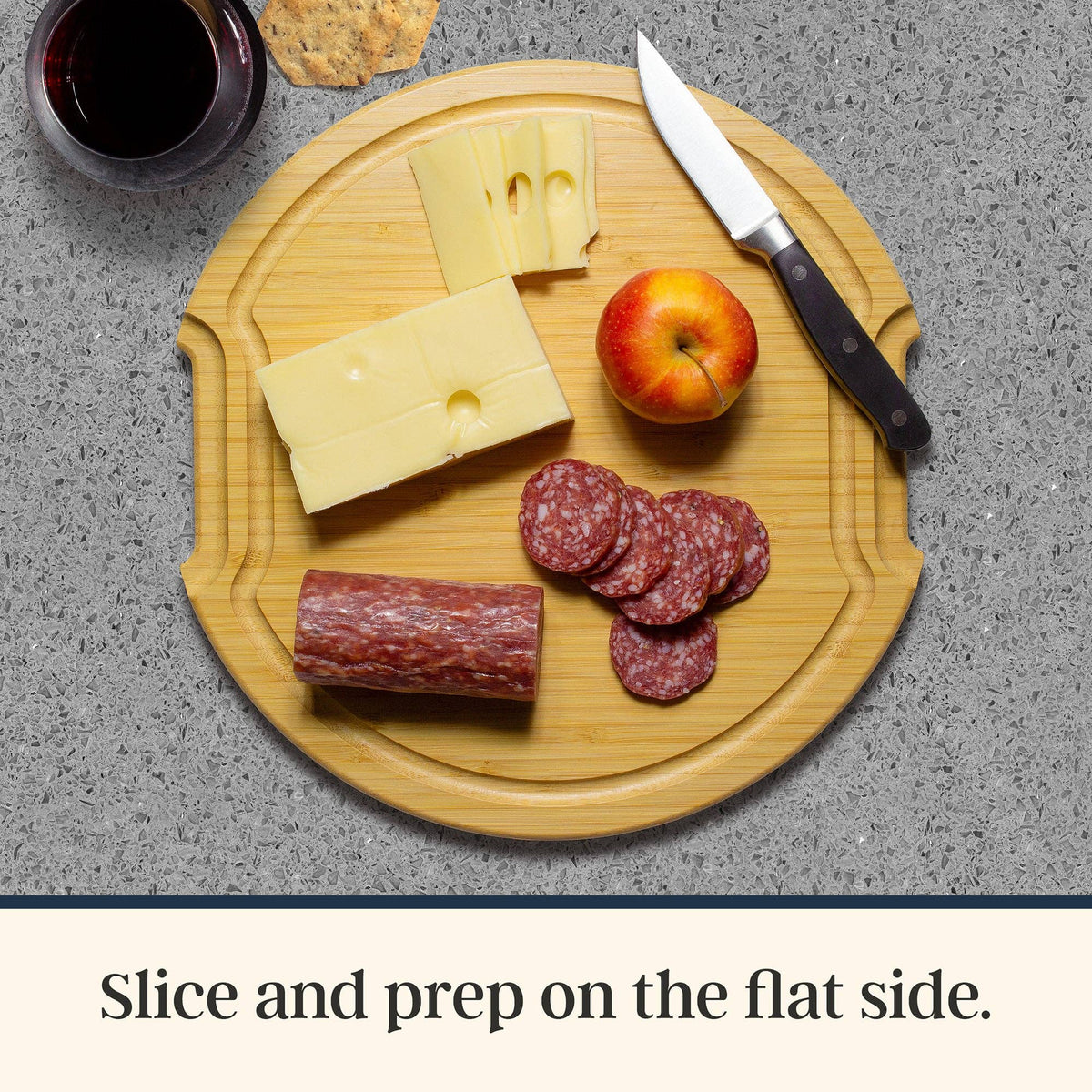 Bamboo charcuterie board featuring sliced cheese, salami, an apple, a knife, a cracker, and a glass of red wine.