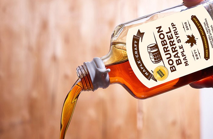 The Bourbon Barrel - Summer Favorites Maple Syrup (6.8 oz) is elegantly poured from a detailed bottle against a wooden backdrop, highlighting its rich bourbon-aged essence, perfect for dessert.