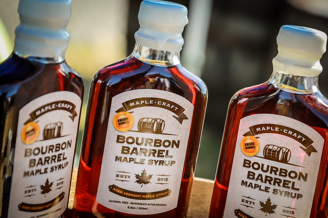 Three bottles of Maple Syrup: Bourbon Barrel - Summer Favorites (6.8 oz), with barrel and maple leaf labels, glisten amber in sunlight, ideal for drizzling on desserts or morning pancakes.