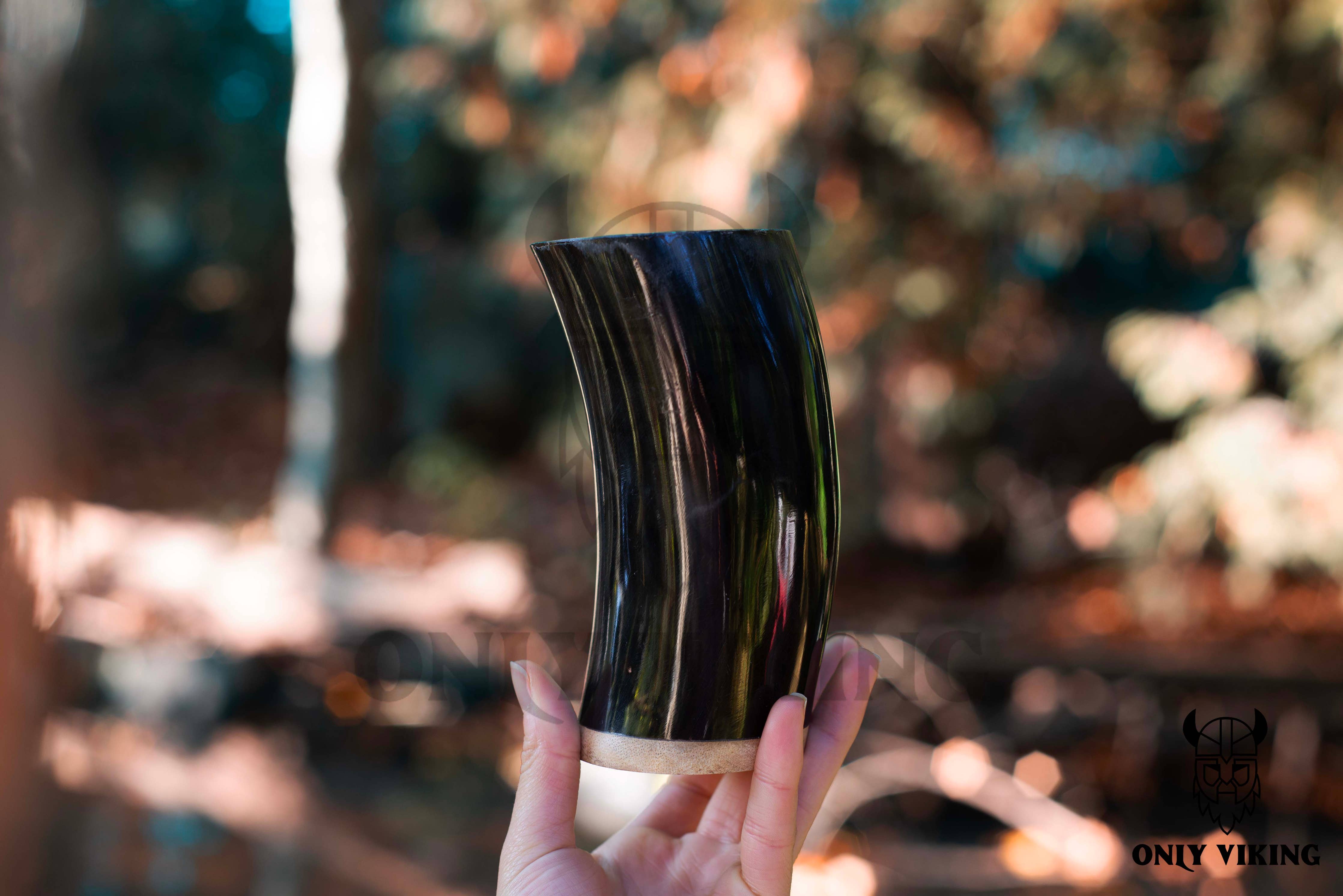 A hand holds the Drinking Horn: Beer Stein 6 Inch with a polished, rounded end outdoors against a blurred background of foliage and sunlight.