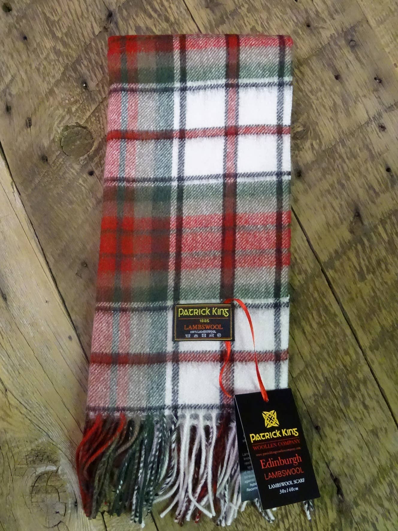 A folded red, green, and white plaid MacDuff Lambswool Scarf with fringe edges rests on a wooden surface, displaying tags labeled Patrick King Lambswool and Edinburgh Lambswool.