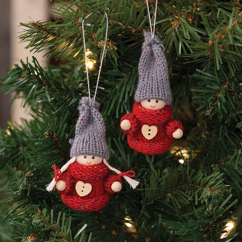 Two gnome ornaments with wooden faces, red sweaters, gray hats, and heart-shaped buttons hanging on a Christmas tree.