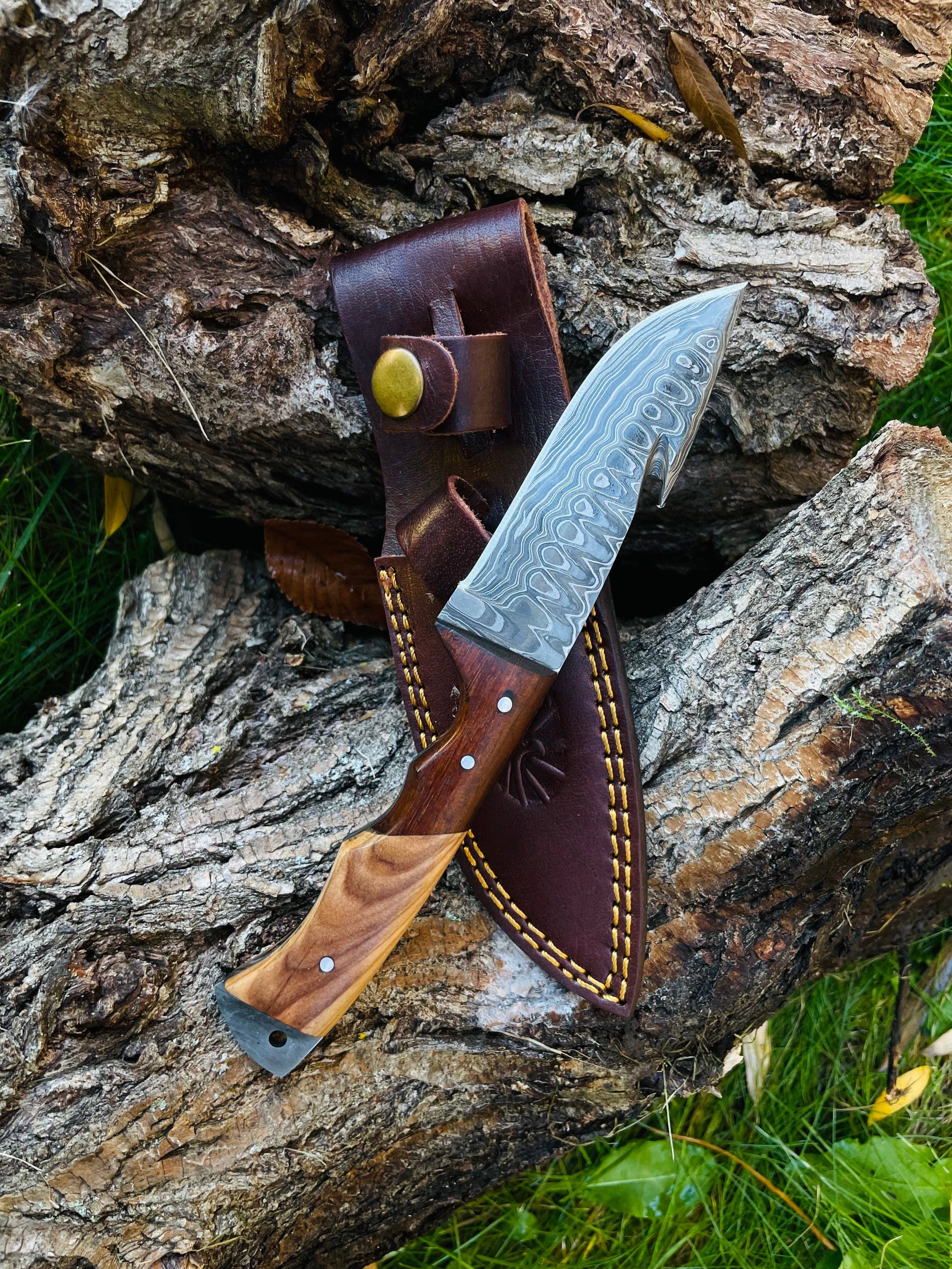 The Knife: Damascus Steel Guthook Skinning Knife, featuring a Damascus steel blade and wooden handle, rests on a brown leather sheath atop rough tree bark.