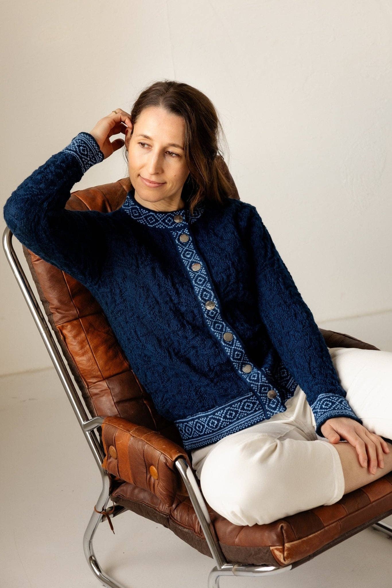 A woman in a Bryggen Women's Norwegian Cardigan (White) and white pants sits in a brown leather chair, looking to the side with her hand touching her hair.