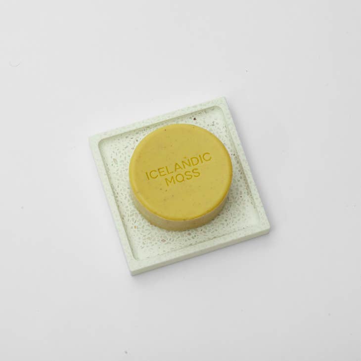 A round yellow soap bar labeled Icelandic Moss sits on an eco-friendly Hello Soap Green Diatomite Soap Dish, against a pristine white background.