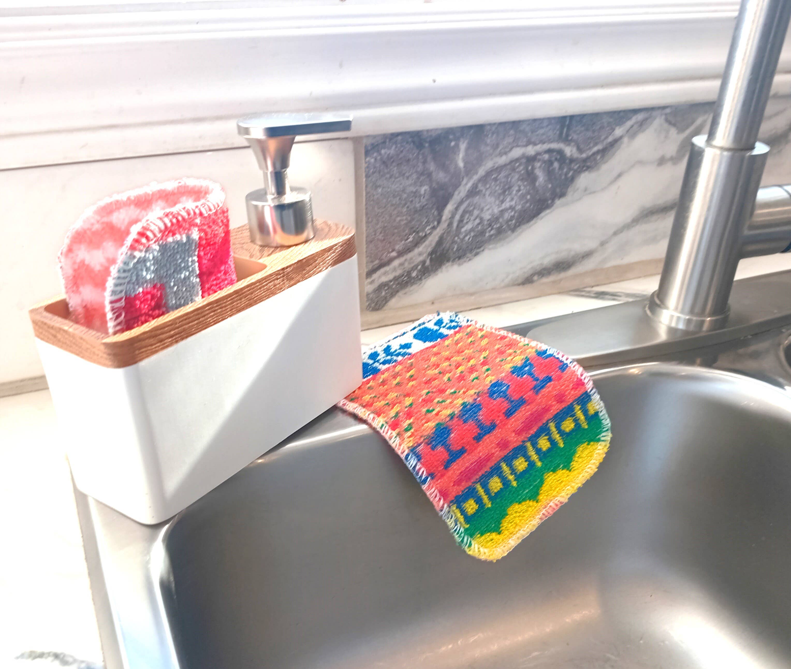 A colorful dishcloth hangs over a stainless steel sink, next to the Scrub and Soap Dispenser—a soap dispenser with a built-in scrubby holder containing two multipurpose scrubbies.