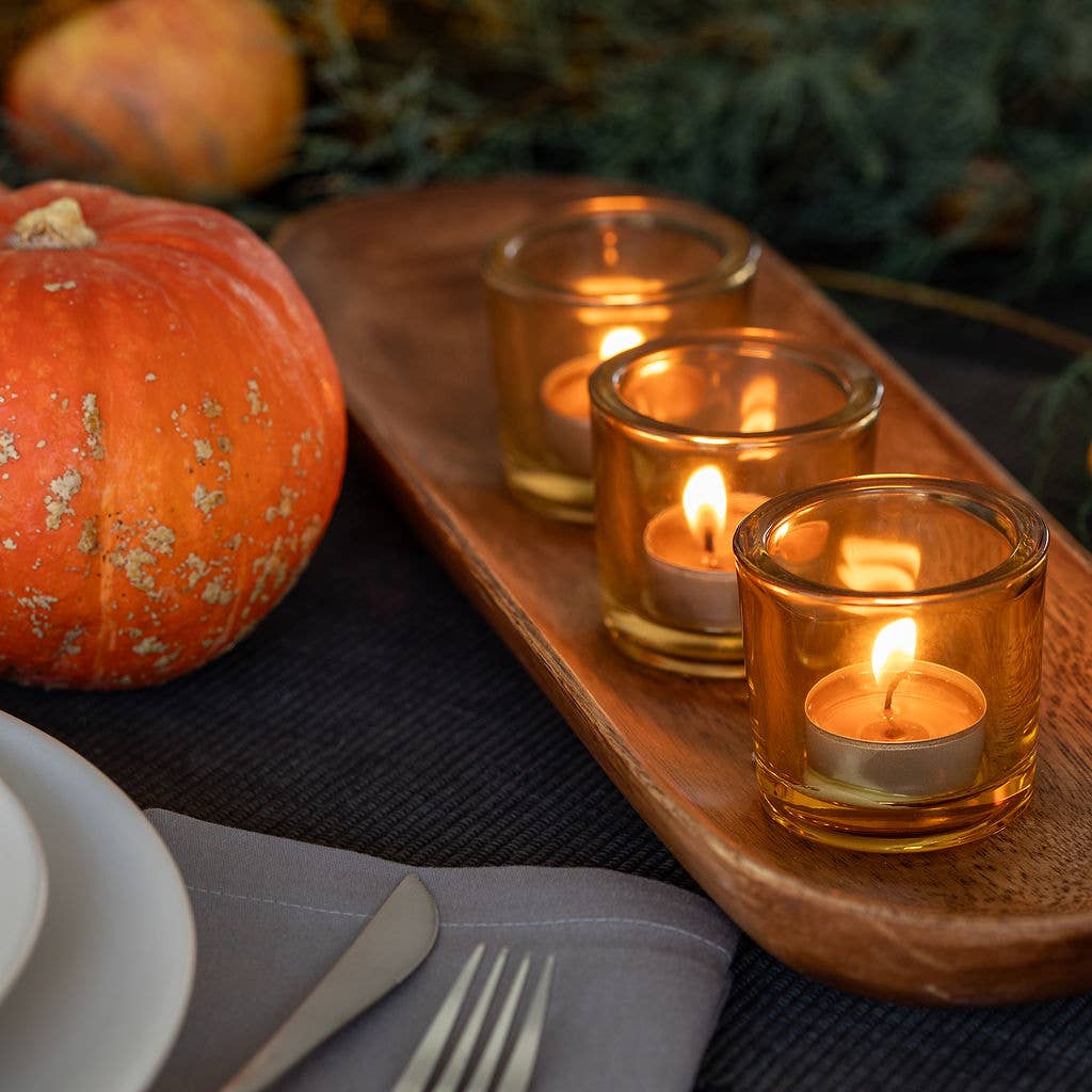 A wooden tray holds three Candle: Pure Beeswax Tea Light Candles – Handmade Raw / Clear Cup in glass holders, accompanied by a pumpkin and dining setup with plates, cutlery, and a napkin.