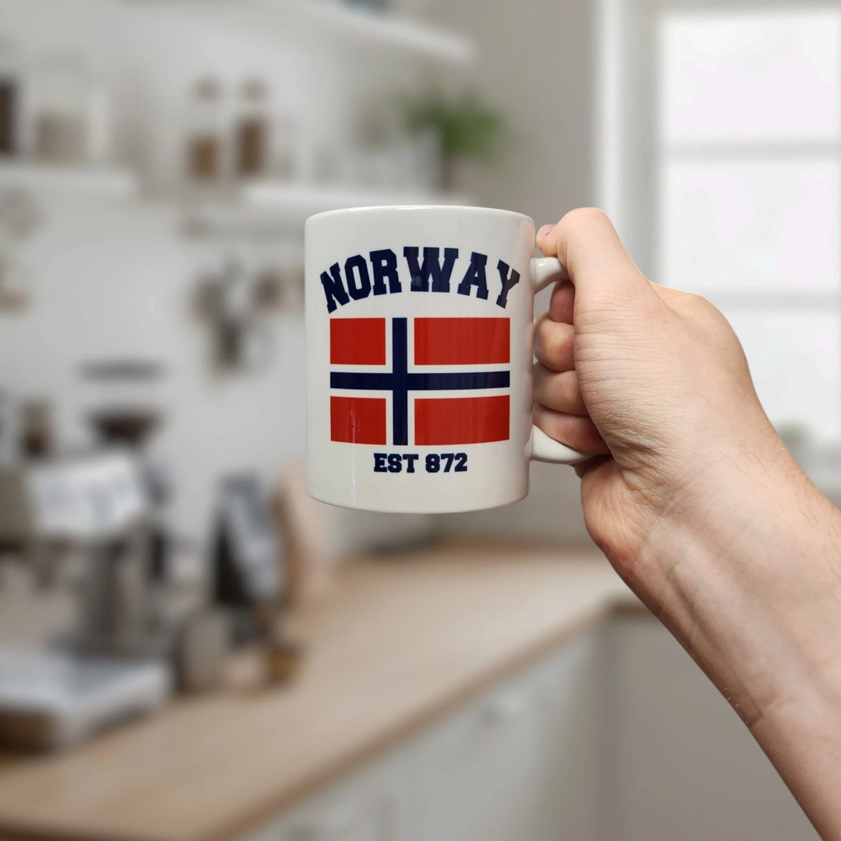A hand holds the Swedish Gift Store Mug: Established Series Norway Flag Mug with Year Founded (11 oz), featuring "NORWAY," the flag, and "EST 872." A blurred kitchen with shelves and a window appears in the background.