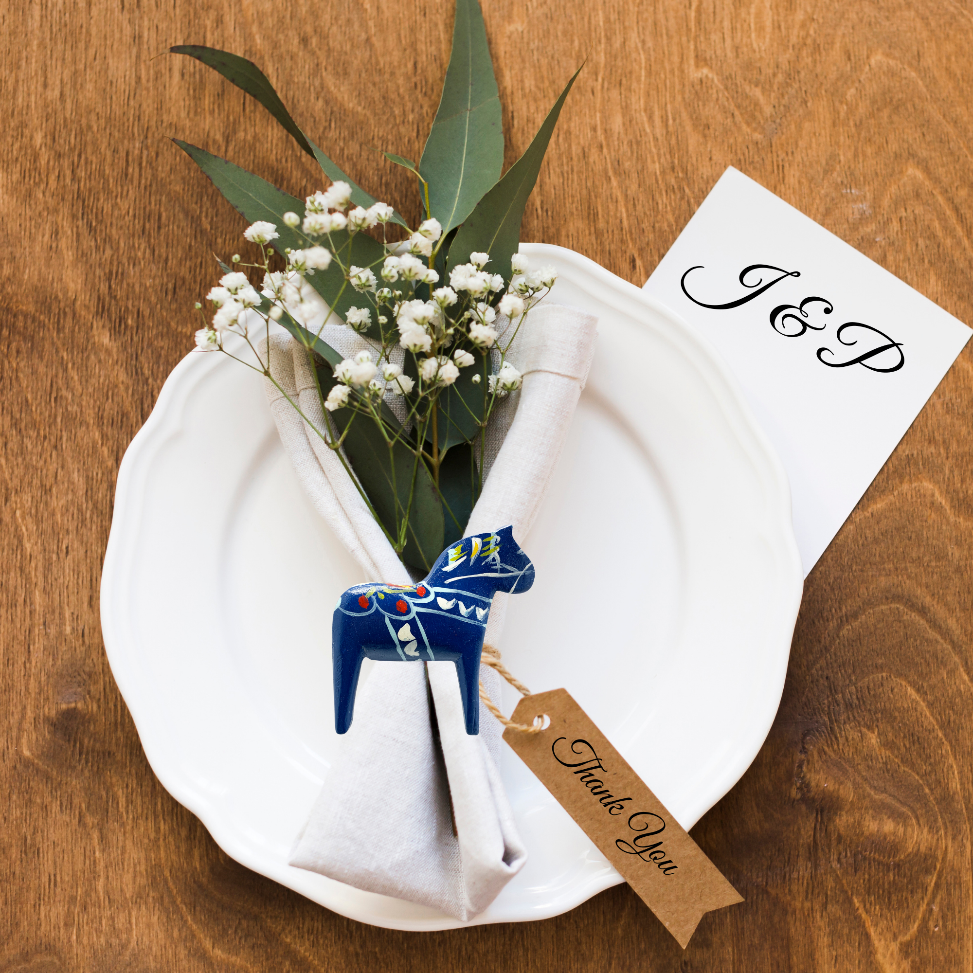 A white plate with a folded napkin, greenery, baby's breath, a Thank You tag, a J & P card, and a blue Swedish Dala Horse Refrigerator Magnet (3”x2.75”) on a wooden table.