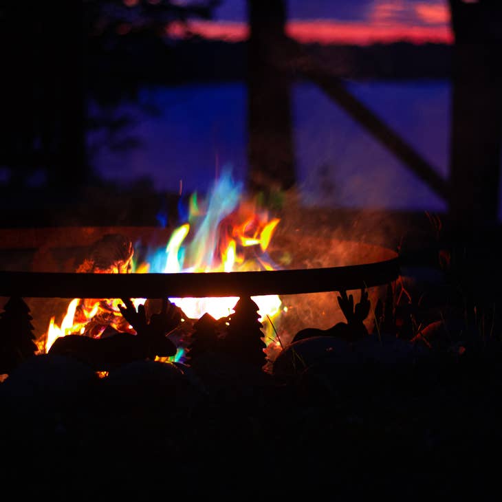 The Fire Color Cones: Northern Lights for Fireside Nights create blue, green, and orange flames in a metal fire ring while a lake and sunset glow in the background.